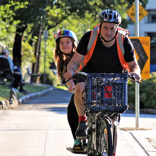 Two people ride a tandem bike along a separated cycle path on a leafy street